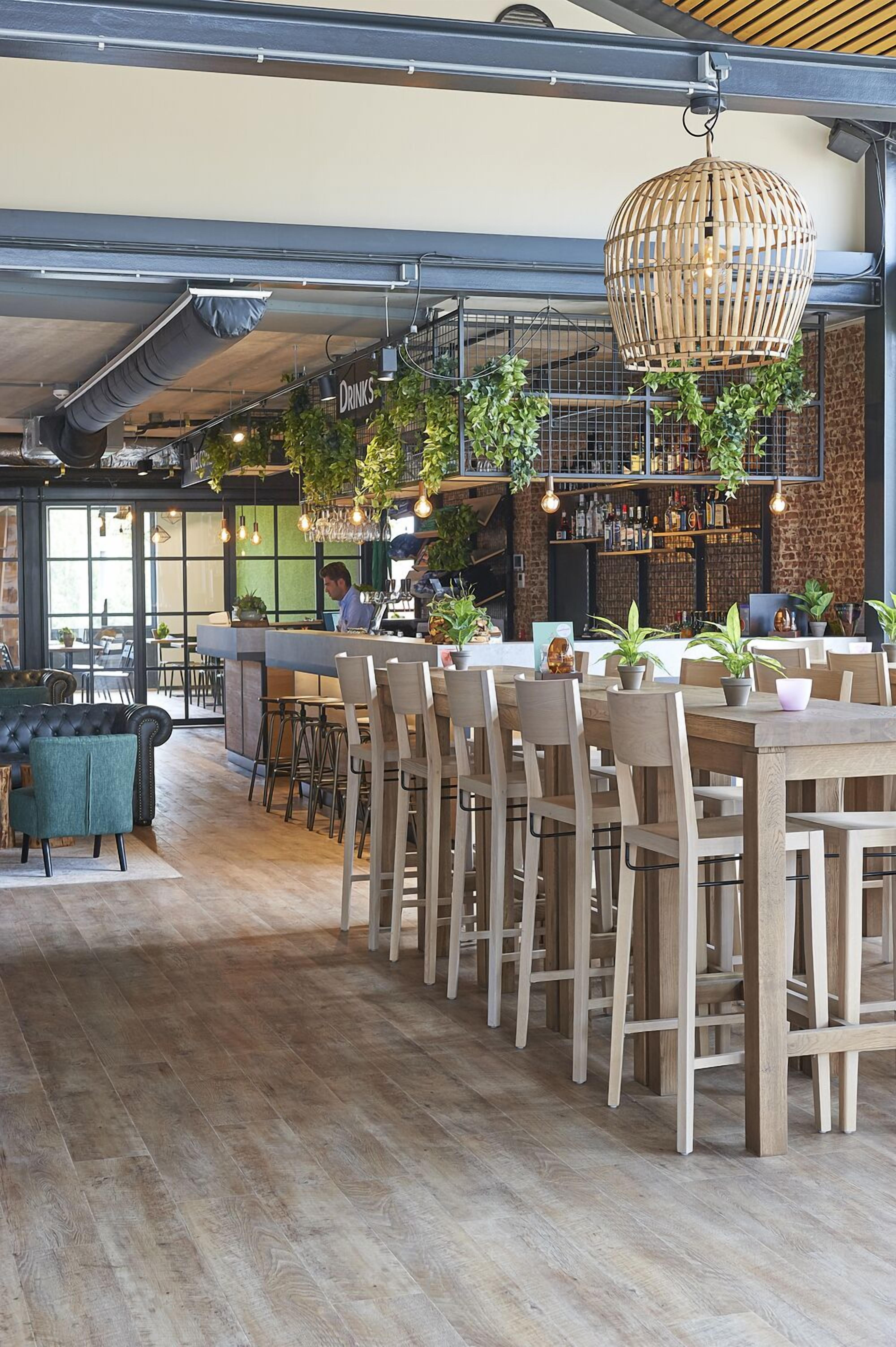 A modern bar area with wooden tables and chairs, hanging light fixtures, and green plants.