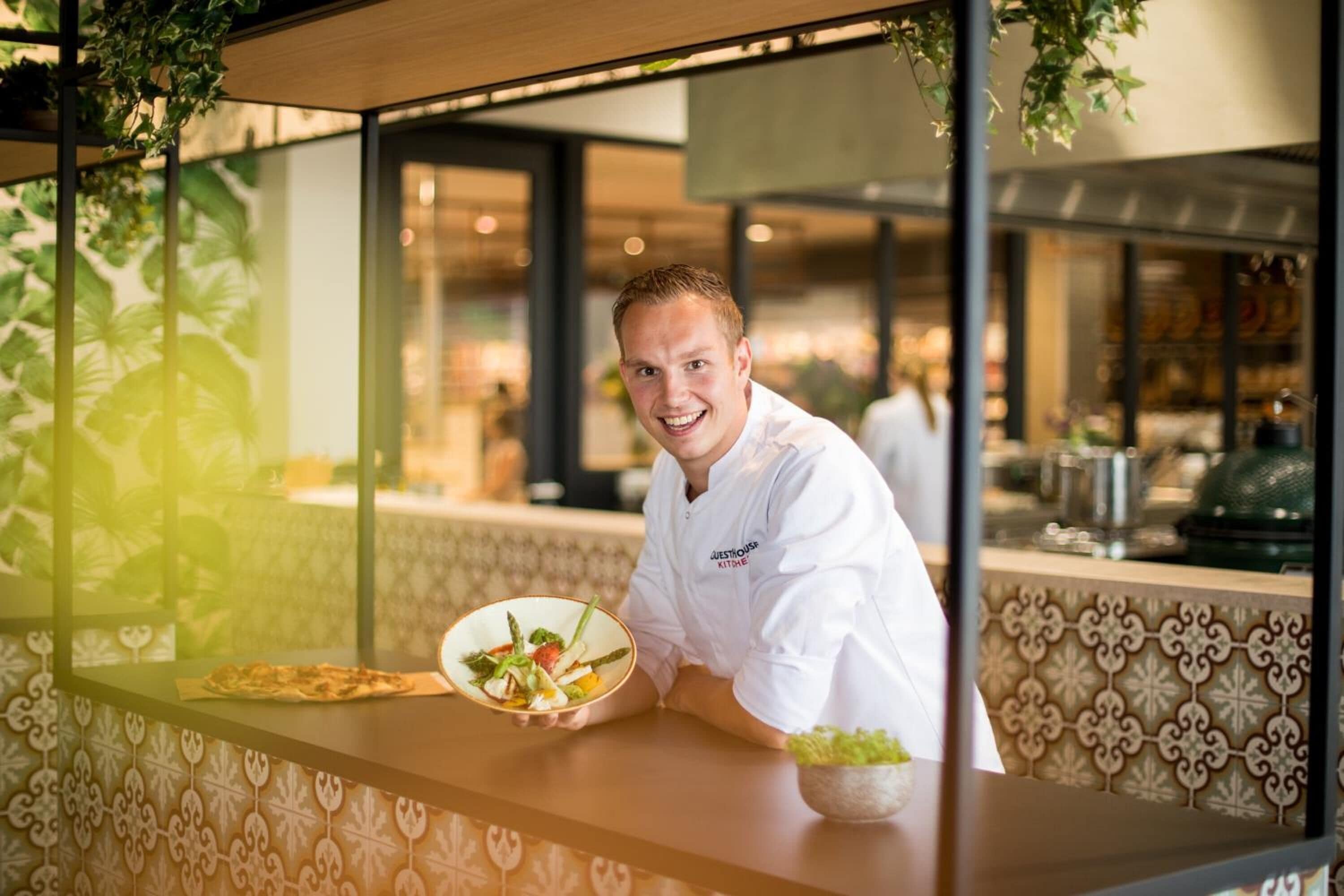 Een kok in een wit uniform met een logo, staand bij een toonbank met een saladeschaal en een bord eten.