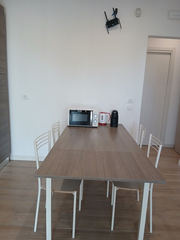 A dining area with a wooden table, white chairs, a microwave, a coffee maker, and a wall-mounted light fixture.