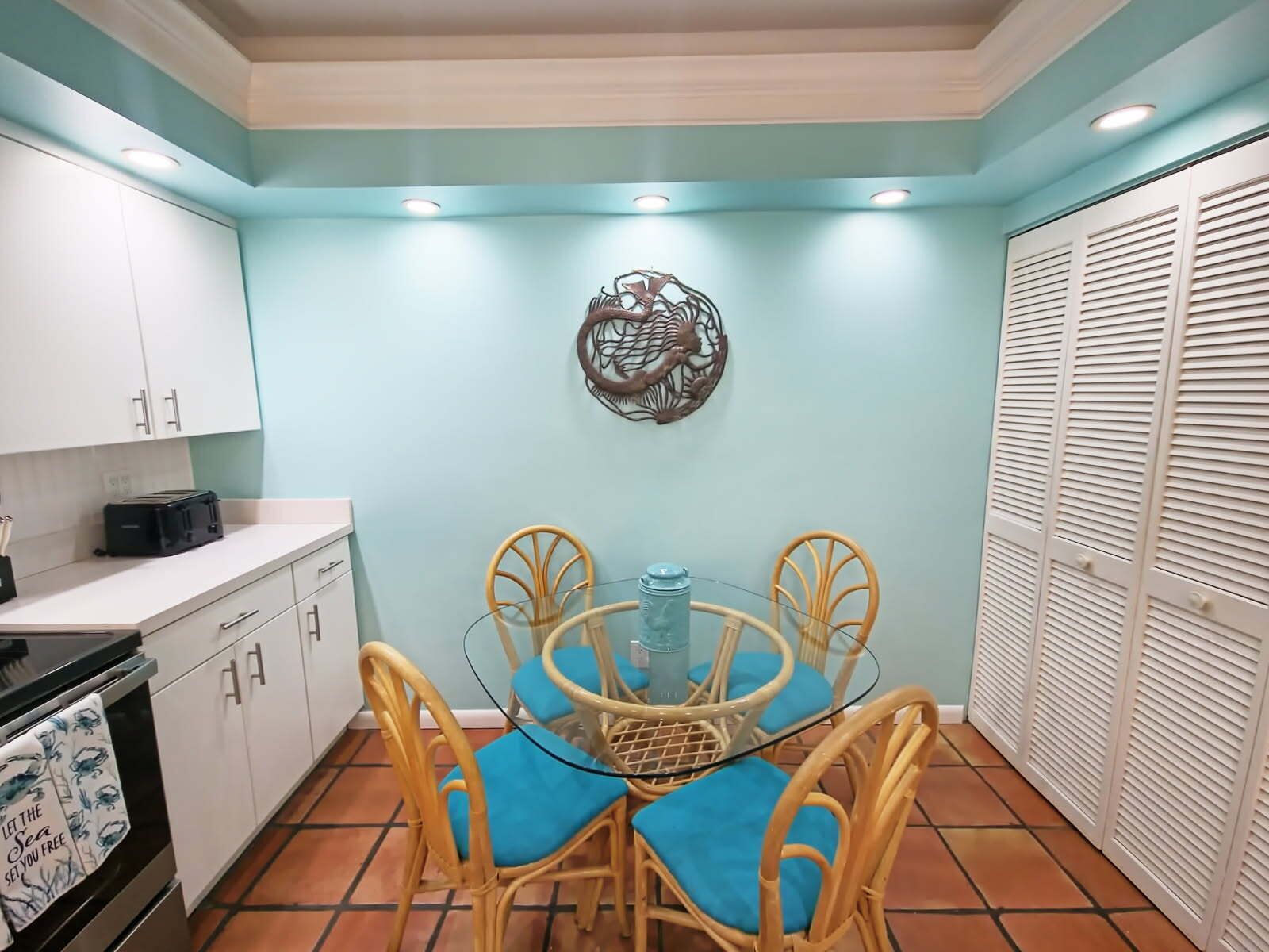 A compact kitchen with white cabinets, a dining table with four chairs, a decorative wall piece, and a white closet with louvered doors.