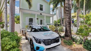 A white Kia SUV parked in front of a light green house with a porch, surrounded by palm trees and greenery. - **CAYO CALYPSO @ HIGGS BEACH** Home / Pool + Last Key White Glove Service... (Key West)