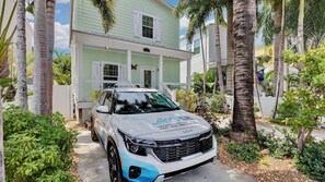A white Kia SUV parked in front of a light green house with a porch, surrounded by palm trees and greenery. - **CAYO CALYPSO @ HIGGS BEACH** Home / Pool + Last Key White Glove Service... (Key West)