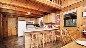 A wooden cabin interior with a kitchen, wooden stools, a dining table, and a window with a view of trees. - 3 king bedrooms with hot tub, pool table, arcade game, great location! (Sevierville)