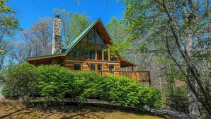 A log cabin with a stone chimney, surrounded by lush greenery and tall trees. - 3 king bedrooms with hot tub, pool table, arcade game, great location! (Sevierville)