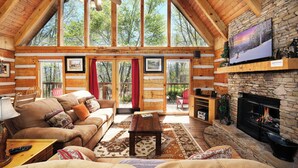 A log cabin interior with a stone fireplace, wooden furniture, and large windows offering a view of the outdoors. - 3 king bedrooms with hot tub, pool table, arcade game, great location! (Sevierville)