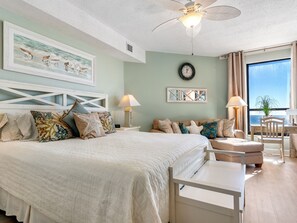 Quarto com cama, mesas de cabeceira, ventoinha no teto, relógio e vista para o mar pela janela.