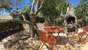 Terrassa exterior amb taula rodona i cadires de fusta, una llar de pedra i vegetació al voltant.
