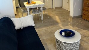 Un saló amb un sofà blau fosc, una taula rodona de centre amb un bol blau, i una porta blanca que dona a una cuina.
