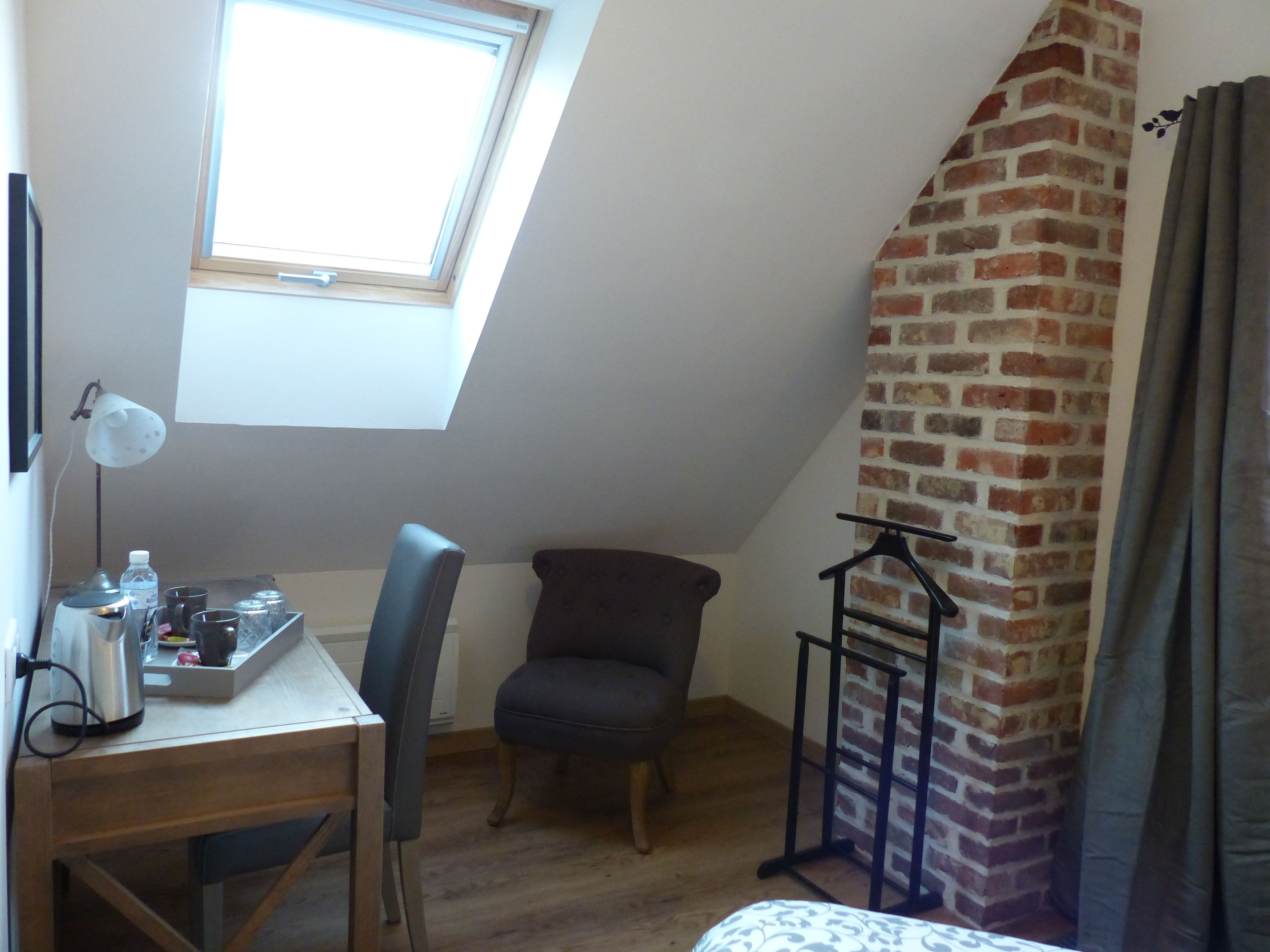 A room with a sloped ceiling, a small table with a kettle, a chair, a hanging coat rack, and a brick wall.