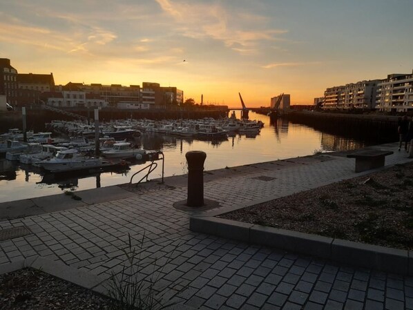 A marina with boats docked, a pedestrian walkway, and buildings in the background during sunset.