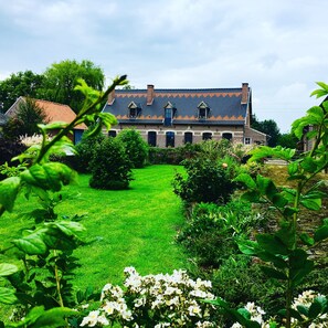 A large, traditional house with a tiled roof, surrounded by a well-maintained garden and a clear sky.