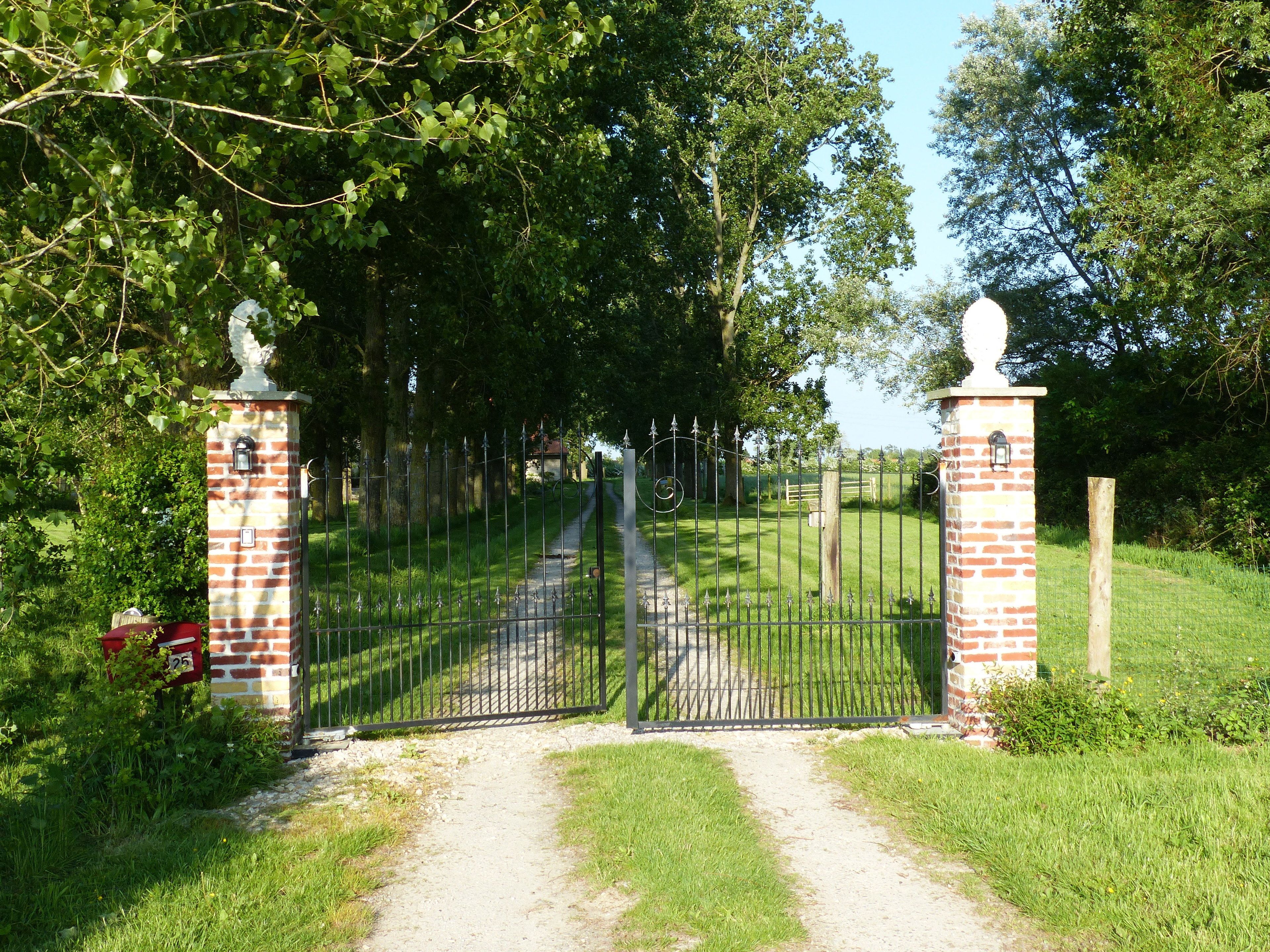 A gated entrance with a brick pillar, a metal gate, and a pathway leading into a green area.