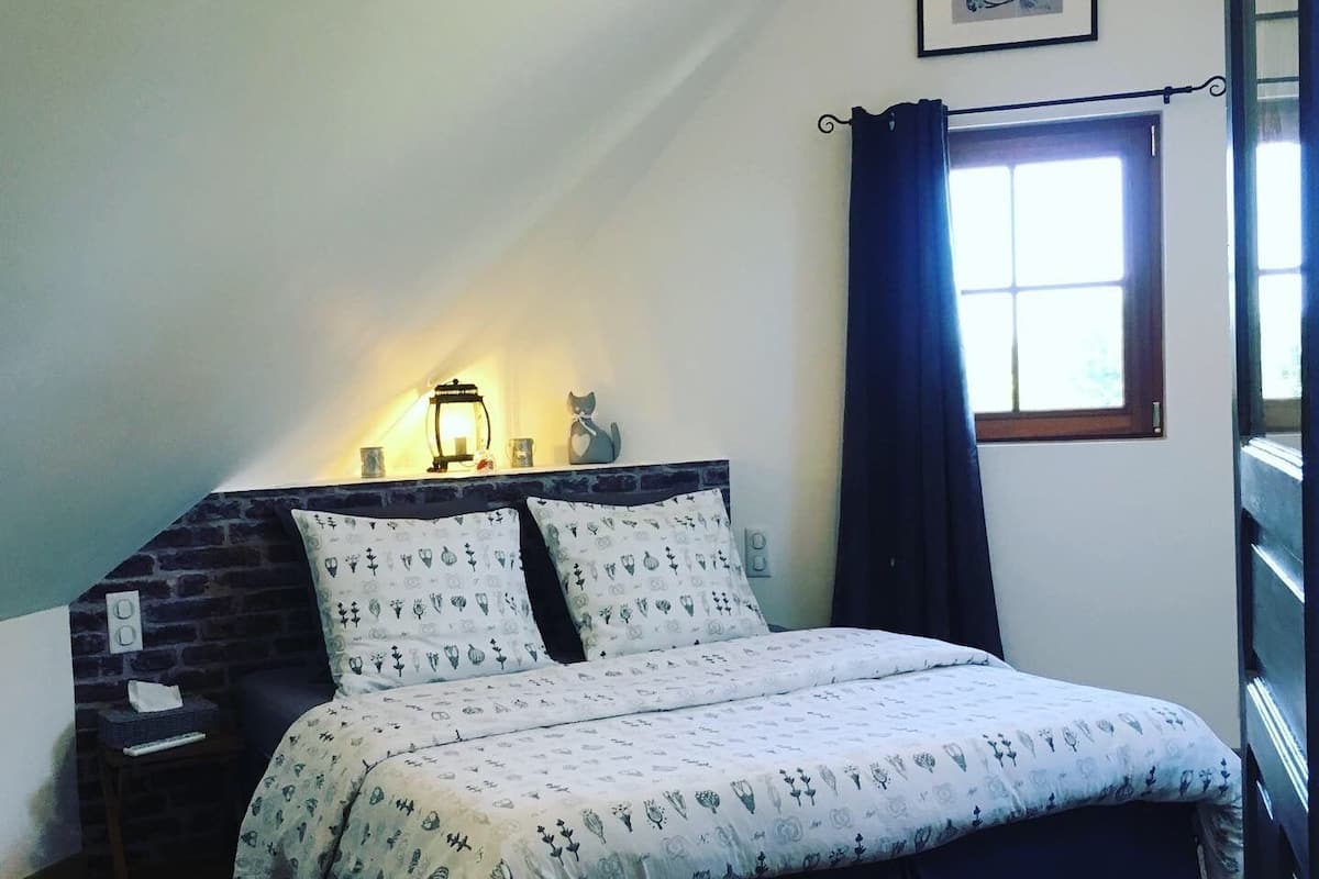 Une chambre à coucher avec un lit, une table de chevet, une lampe, une fenêtre avec des rideaux et un tableau encadré au mur.