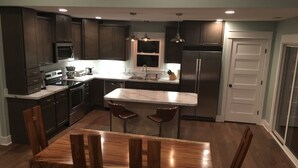 A modern kitchen with dark cabinetry, stainless steel appliances, a marble island, and a wooden dining table with chairs. - The Weathervane | 3BR Carova Beach 4x4 OBX | 4 night stay available | Pets (Carova)