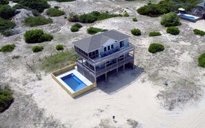 Una casa sulla spiaggia a due piani con piscina, circondata da sabbia e vegetazione rada.