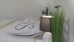 Une chambre à coucher bien rangée, avec un lit, une lampe de chevet, un vase décoratif contenant des plantes vertes et un oreiller orné d’une grande lettre.