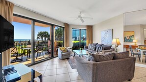 Een ruime woonkamer met een bank, salontafel en televisie. Het heeft een balkon met buitenmeubilair en uitzicht op palmbomen.