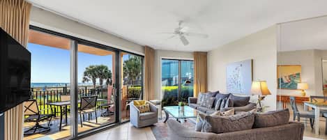 A spacious living area with a sofa, coffee table, and TV. It features a balcony with outdoor furniture and a view of palm trees.