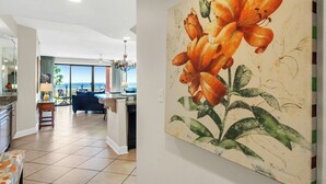 Een ruime woonkamer met een groot schilderij van bloemen aan de muur, een uitzicht naar buiten en een moderne keuken op de achtergrond.