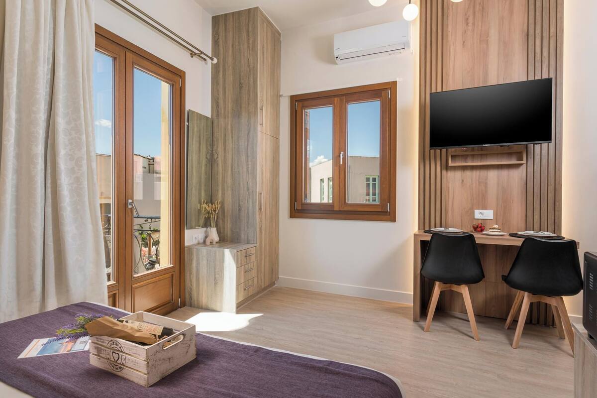 A modern bedroom with a large window, a wooden wardrobe, a desk with two chairs, and a flat-screen TV mounted on the wall.