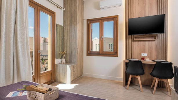 A modern bedroom with a large window, a wooden wardrobe, a desk with two chairs, and a flat-screen TV mounted on the wall. - Porto Orion Studios (Chania)