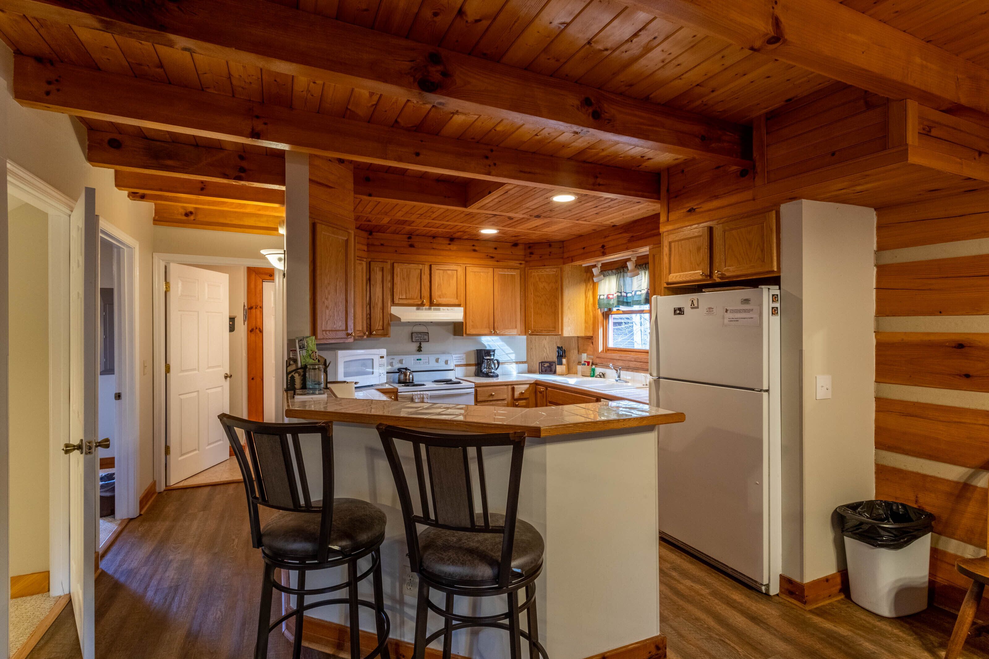 A kitchen with wooden cabinets, a white refrigerator, a microwave, and a sink. There are two bar stools and a trash can.