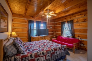 Une chambre confortable dans un chalet, avec un intérieur en bois, un lit recouvert d’une courtepointe, un canapé rouge, un téléviseur posé sur une commode en bois et un ventilateur de plafond.