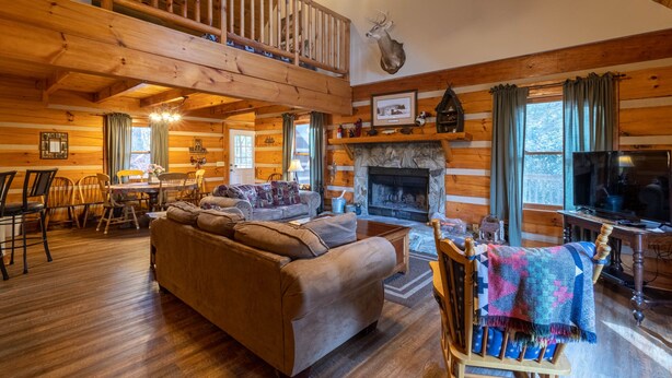 A spacious log cabin interior with a high ceiling, a fireplace, a dining area, and a cozy living room with a sofa and a television.