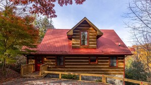 A log cabin with a red roof, wooden exterior, and a small porch with a railing.