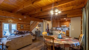 A cozy cabin interior with a wooden ceiling, a dining area with a quilt-covered table, and a living room with a sofa and a staircase leading upstairs.