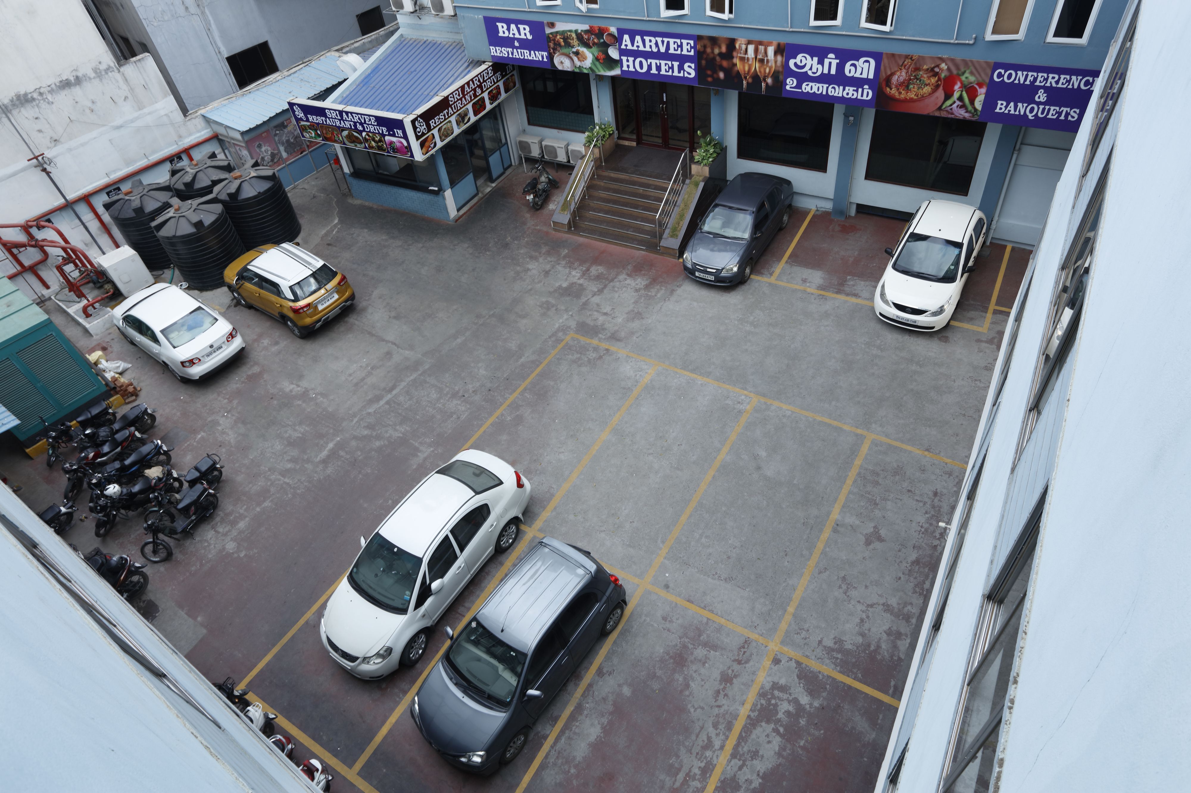 Ein Parkplatz mit mehreren Autos und Motorrädern, eine Treppe zu einem Gebäude mit Schildern für eine Bar und ein Restaurant, sowie ein Hotel.