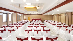 Un salón de conferencias con filas de sillas blancas adornadas con listones rojos, un escenario con mantel rojo y un revestimiento de pared de madera.
