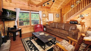 A cozy cabin interior with a wooden interior, a brown leather sofa, a red chair, a black coffee table, and a staircase leading to an upper level. - Pet friendly 2 bedroom with hot tub, Smoky Mountain view, and arcade game! (Sevierville)