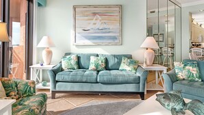 Een woonkamer met een blauwgroene bank, bloemenkussens, een schilderij van een zeilboot en een sculptuur van een vis.
