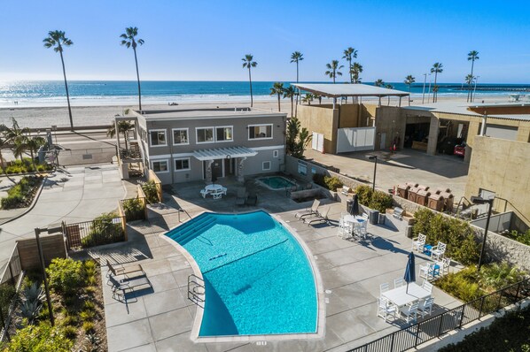 A pool area with a clear view of the ocean, a building with a patio, and a beach in the background.