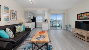 A modern living room with a sofa, coffee table, and TV stand. There is a kitchen area with cabinets and a dining table. A bicycle is visible in the background. - 303B Perfect Location  Beach Across the Street - Walk to Shops Dining - Harbor Views (Oceanside)