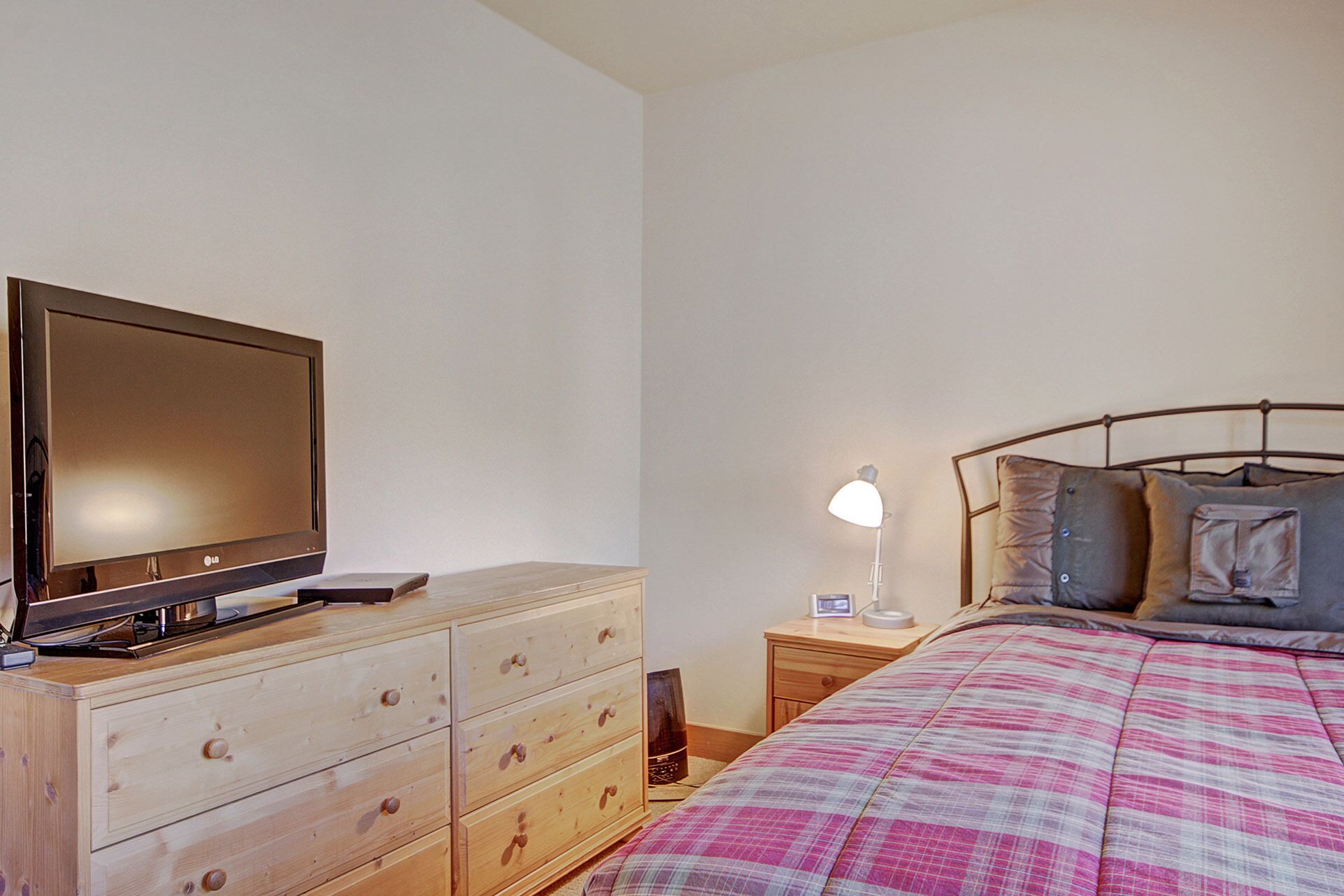 A bedroom with a wooden dresser, a bed with a plaid comforter, a bedside lamp, and a television on the dresser.