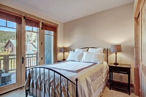 A bedroom with a bed, two bedside tables with lamps, a large window with blinds, and a view of a house outside.