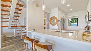 A modern kitchen with a central island, bar stools, a hanging light fixture, and a spiral staircase. - Brand New Kitchen & Updated Living Space - Oceanfront #216-2 (Lahaina)