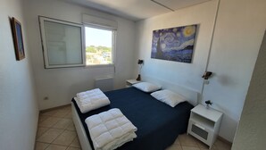 Un dormitorio con cama, mesita de noche, lámpara de noche, un cuadro y una ventana.
