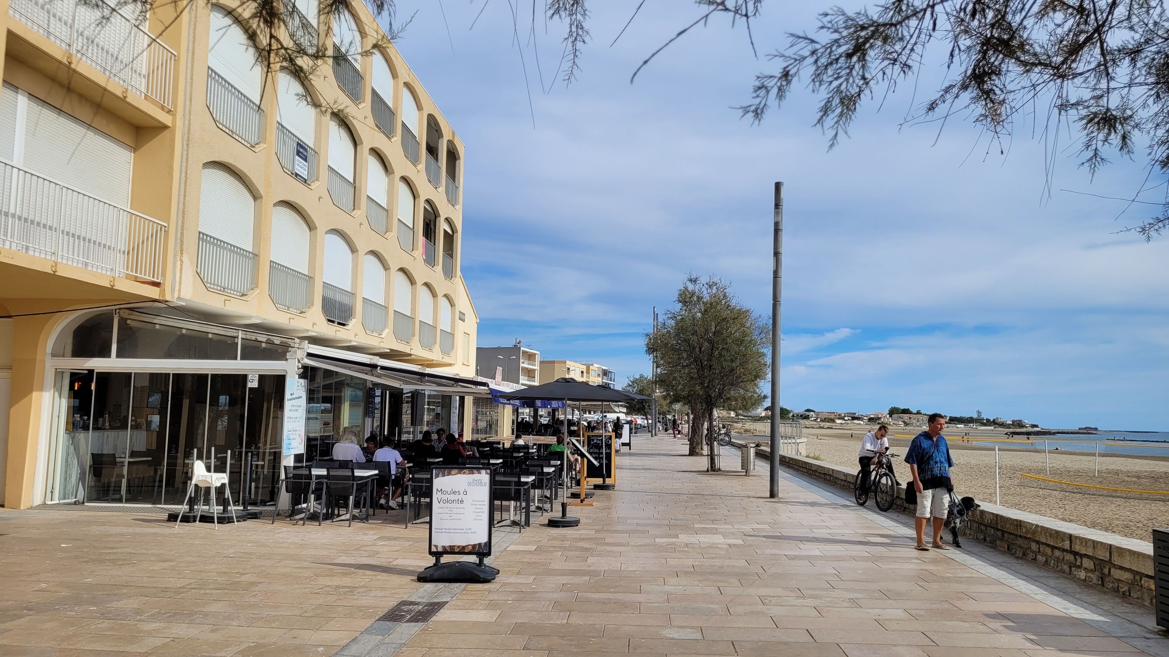 A seaside promenade with outdoor seating at cafes, a beach in the background, and a building with balconies.