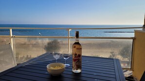 Una mesa con una botella, dos vasos y un bol sobre un balcón con vista al mar.