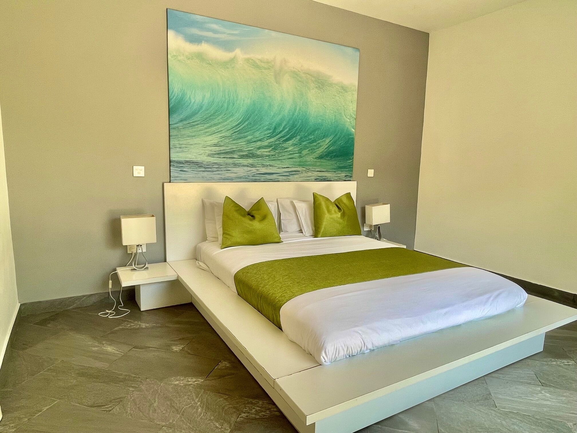 A modern hotel room with a large bed, green pillows, a white headboard, and a painting of a wave on the wall.