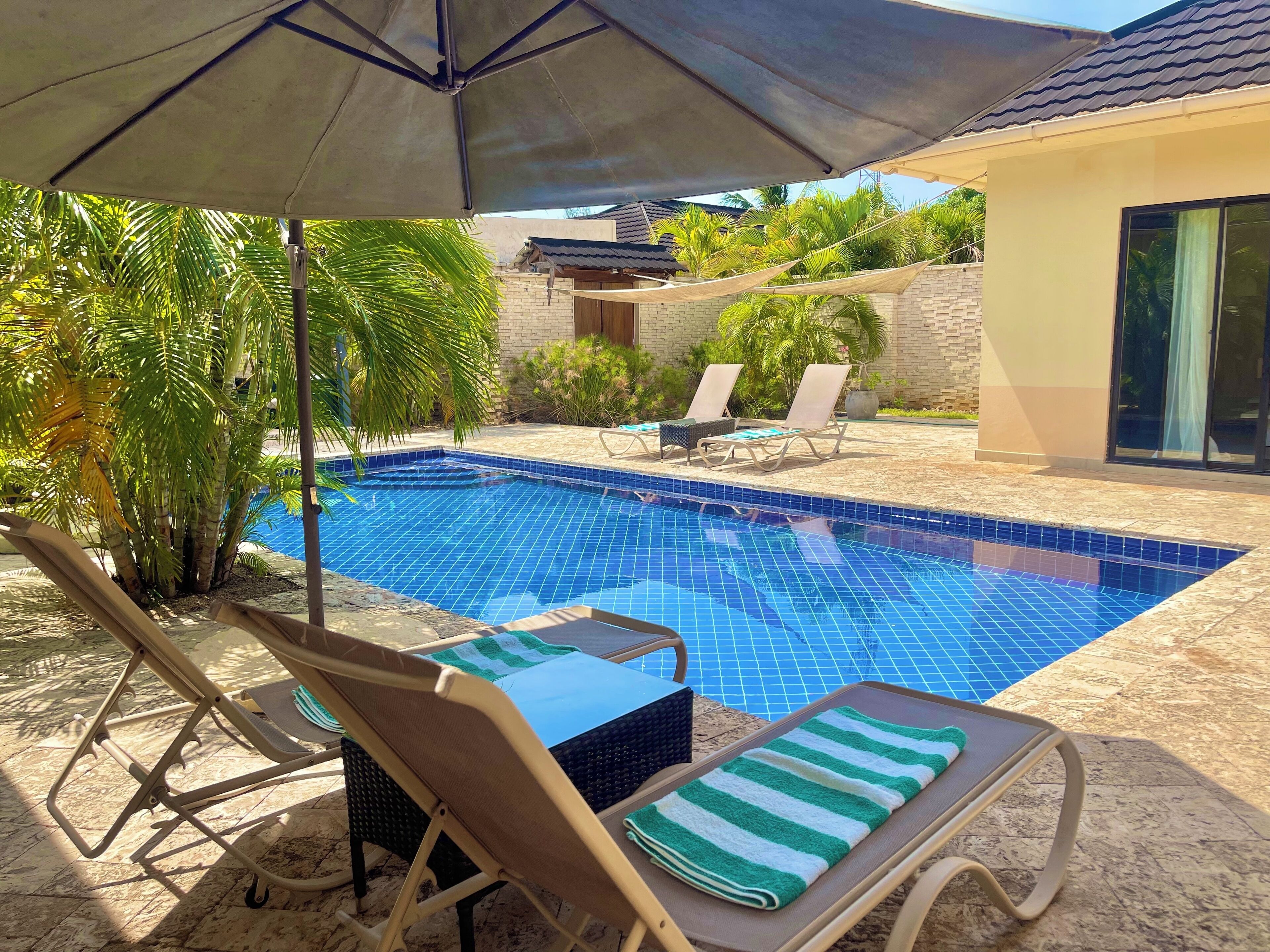 A swimming pool with lounge chairs and an umbrella, surrounded by greenery and a building.