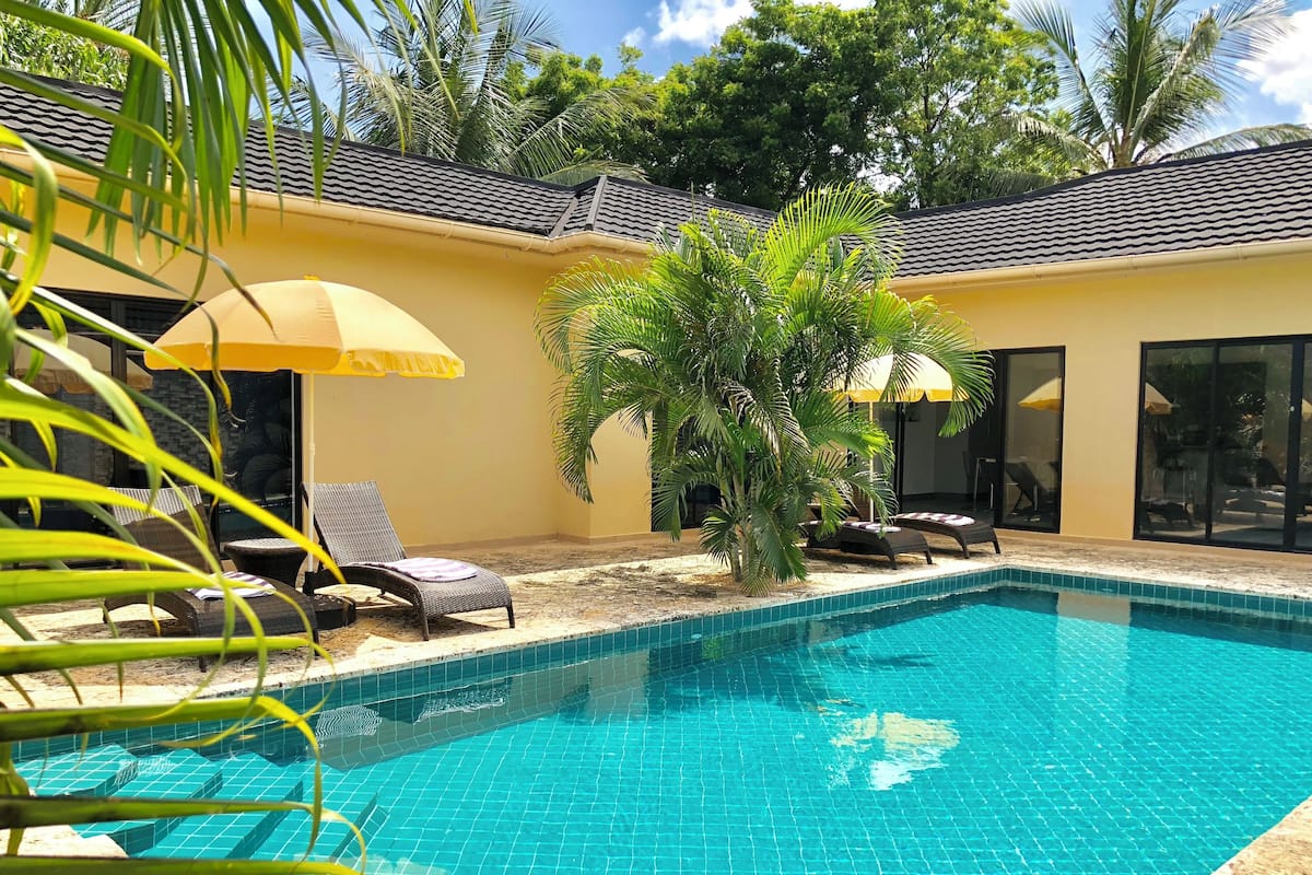 A swimming pool with tiled flooring, surrounded by lounge chairs and umbrellas, with palm trees and a building in the background.