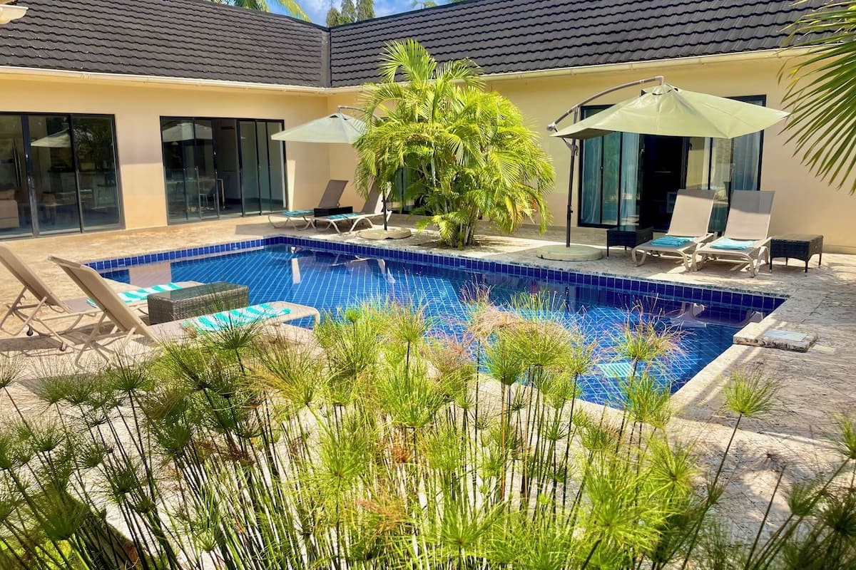 A swimming pool with lounge chairs and umbrellas, surrounded by tropical plants and a modern building with large windows.