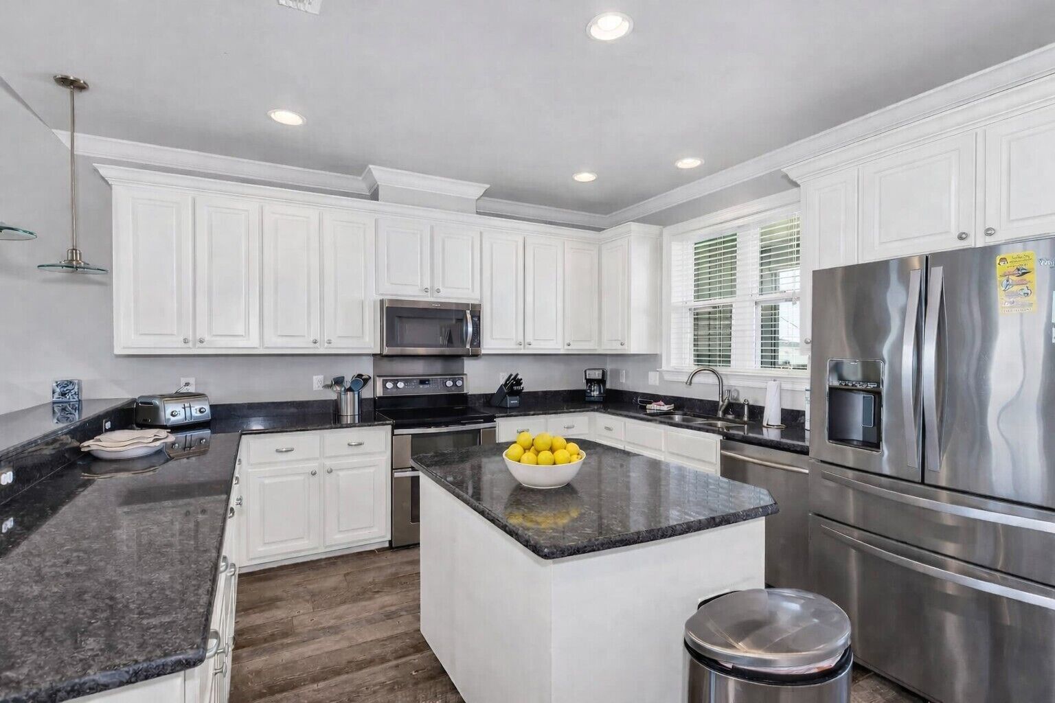 A modern kitchen with white cabinets, a central island with a bowl of lemons, stainless steel appliances, and a black granite countertop.