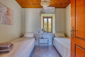 Quarto com duas camas de solteiro, uma mesa pequena, uma luminária e uma janela com vista para o verde.