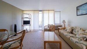 A living room with a sofa, a coffee table, a TV, and a chair. There is a window with curtains, a lamp, and a picture on the wall. - Turtle Bay Guava.Price Drop September start at $200 nightly. (Kahuku)