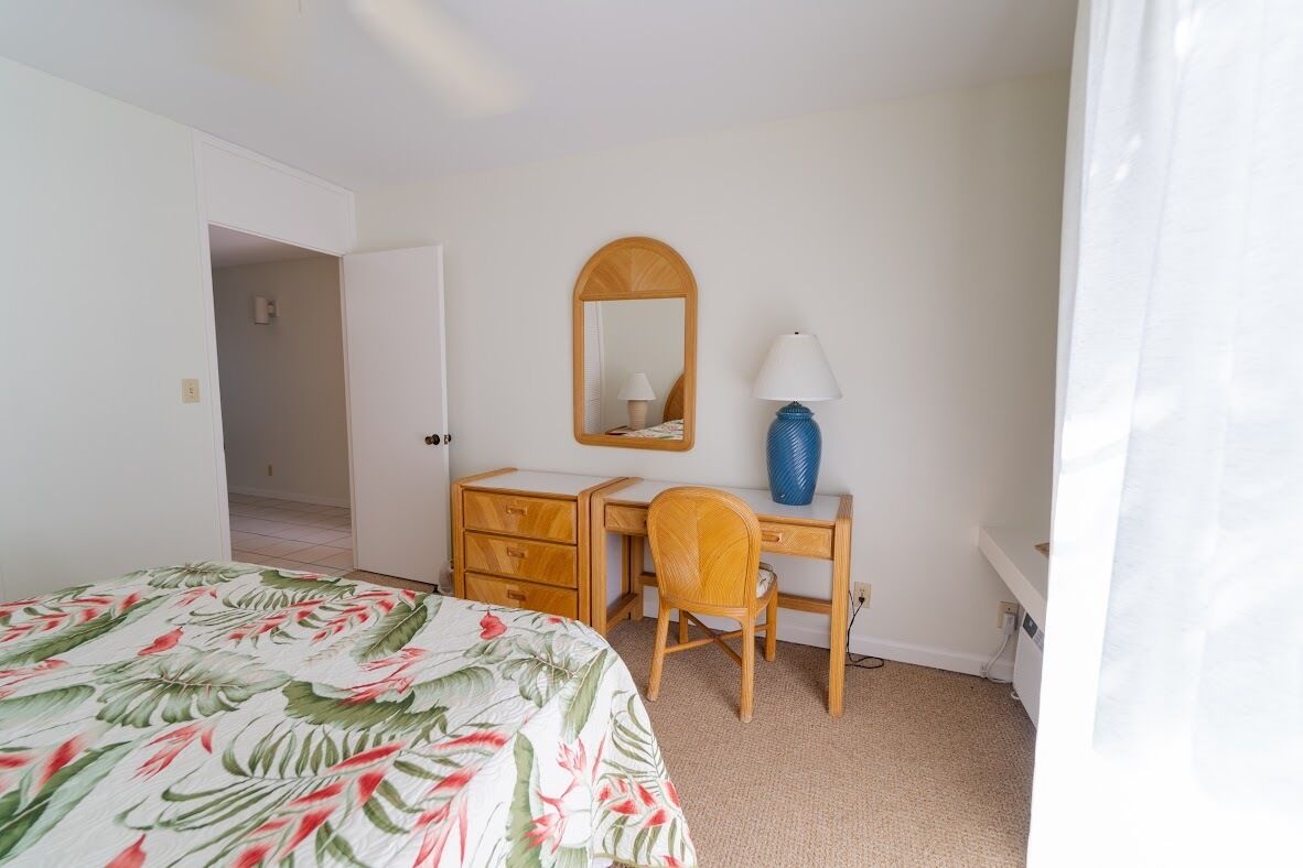 A bedroom with a bed, a wooden desk with a chair, a mirror, and a lamp.